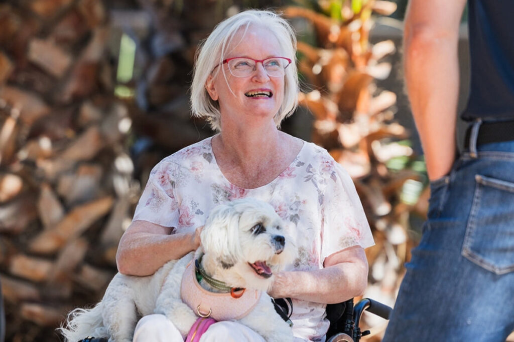 Woman with cerebral palsy sitting in a wheelchair holding her small white dog Willow, smiling outdoors in a garden setting